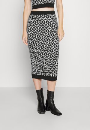 Woman wearing a black and white geometric patterned midi skirt, black crop top, and black ankle boots, standing against a plain background.