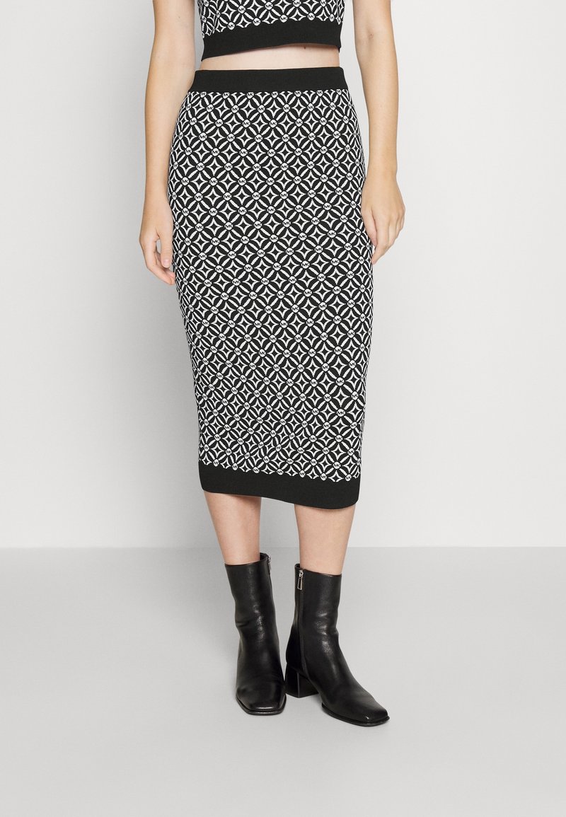 Femme portant une jupe midi à motif géométrique noir et blanc, un crop top noir et des bottines noires, debout sur un fond uni.