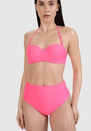 Kobieta ubrana w jasnoróżowy, bezramiączkowy top od bikini z ramiączkami na szyję i wysokim stanem bikini na jednolitym tle.