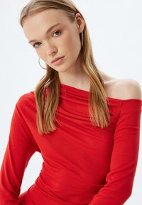 Top rosso scollato con maniche lunghe, caratterizzato da un dettaglio di tessuto arricciato attorno al collo. Materiale in cotone leggero e liscio.