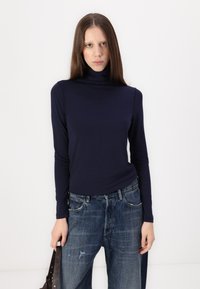 Mornarsko modra dolga majica s turtleneck ovratnikom, izdelana iz mehkega materiala, v kombinaciji z raztrganimi, ohlapnimi modrimi kavbojkami. Model drži temno torbo.
