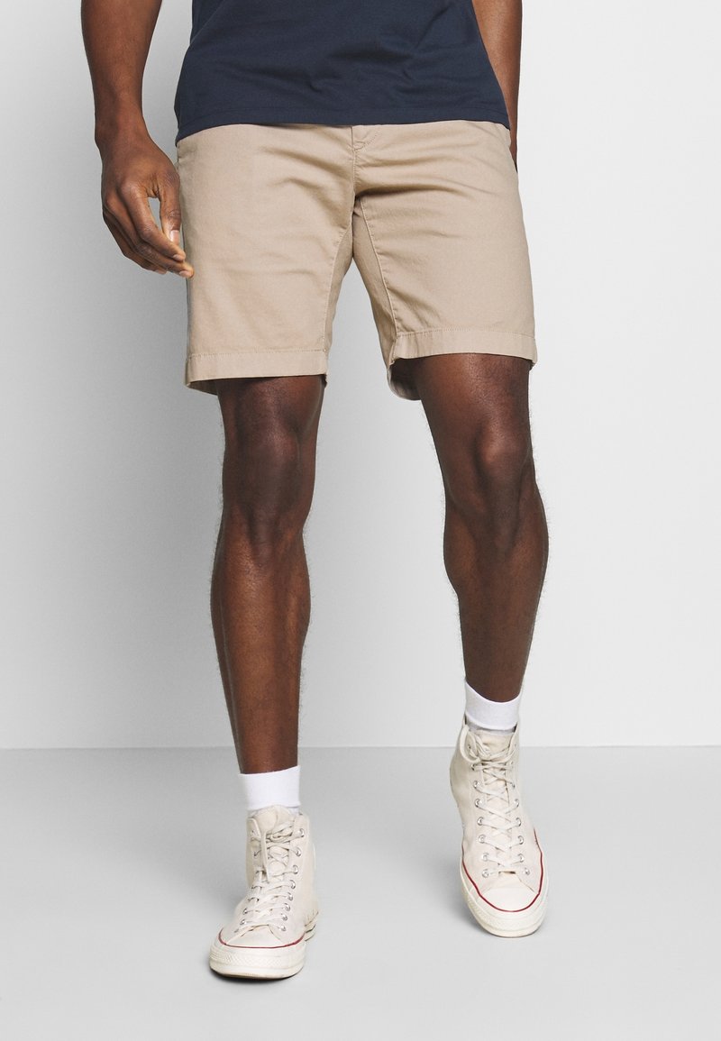 Homme portant un short beige à hauteur des genoux, des baskets blanches montantes et des chaussettes blanches, debout devant un fond neutre.