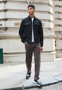 Schwarze Wildlederjacke mit zwei Fronttaschen, einem hellgrauen Pullover darunter und braunen Hosen. Schwarze Sneakers mit weißen Akzenten runden das Outfit ab.