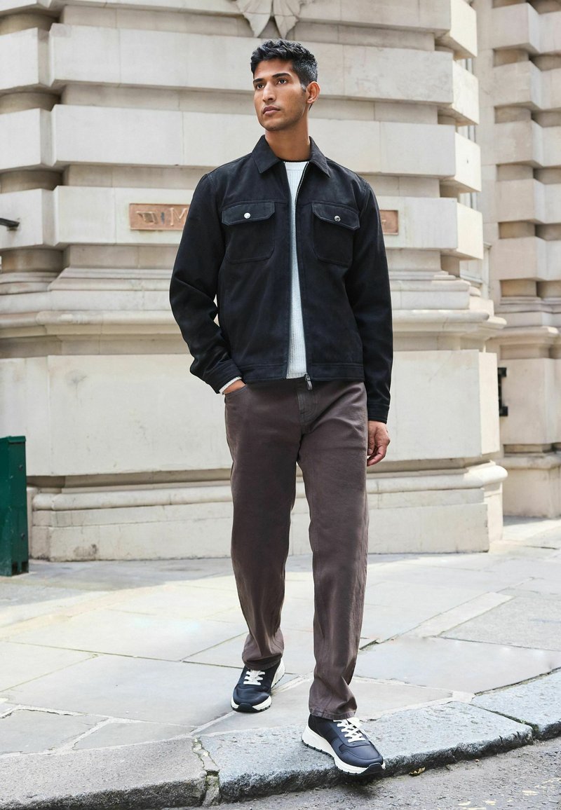 Schwarze Wildlederjacke mit zwei Fronttaschen, einem hellgrauen Pullover darunter und braunen Hosen. Schwarze Sneakers mit weißen Akzenten runden das Outfit ab.