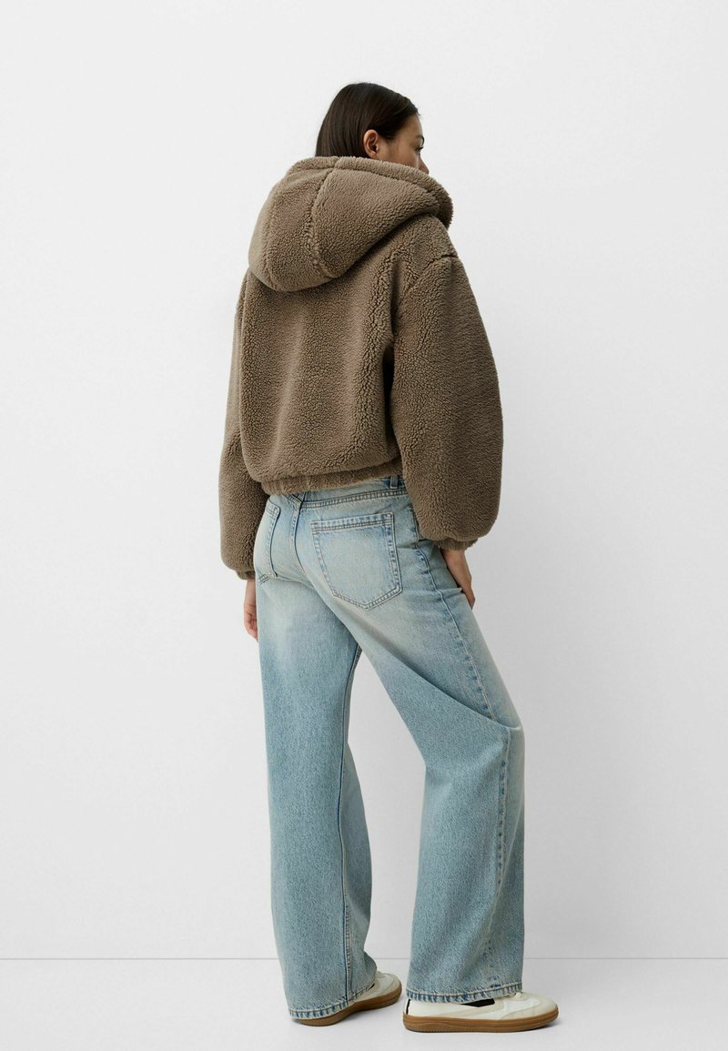 Veste à capuche en polaire marron au design court, associée à un jean large bleu clair et des baskets blanches. Matériaux lisses et texturés.