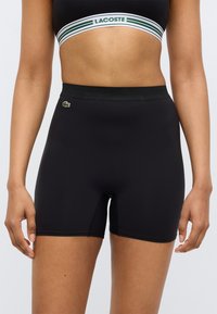 Shorts de sport noirs en tissu extensible, avec un design uni, une taille ajustée et un petit logo Lacoste vert sur le côté gauche.