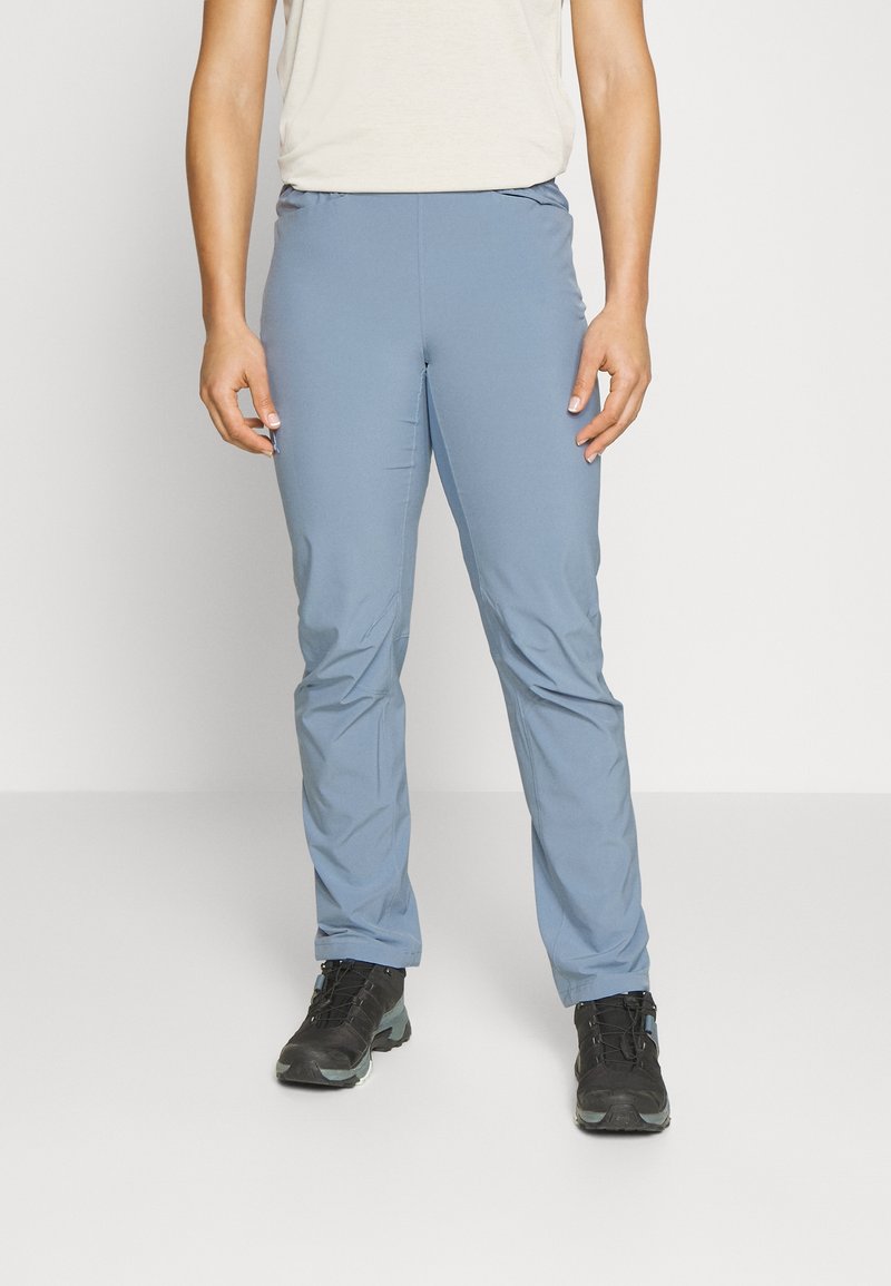 Personne portant un pantalon slim bleu clair et des chaussures de randonnée noires, debout devant un fond blanc uni.