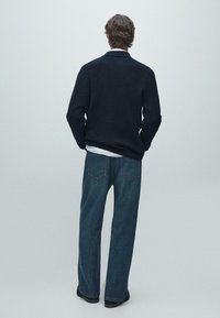 Cardigan blu navy a coste, vestibilità casual, abbinato a jeans scuri larghi in denim. Dettagli visibili includono tasche posteriori e camicia bianca semplice sotto.