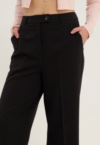 Pantalon noir sur mesure avec une texture lisse, doté d'une fermeture à un bouton et de poches latérales. Coupe droite classique.