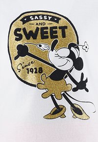 Camiseta blanca con un gráfico de un personaje de dibujos animados con un diseño en negro y dorado, texto "SASSY AND SWEET" y "Desde 1928", y un acento de flor.