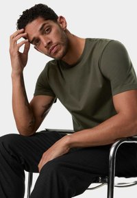 Kortærmet olivengrøn t-shirt lavet af glat stof, med en rund halsudskæring. Kombineret med sorte bukser, siddende på en stol.