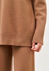 Pulover tricotat maro cu mansete și tiv cu nervuri, purtat peste pantaloni tricotati maro asortați. Textură moale, design simplu.