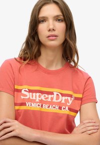 Ung kvinde med lysebrunt hår iført en koralfarvet SuperDry Venice Beach CA T-shirt, armene krydsede, foran en ensartet hvid baggrund.