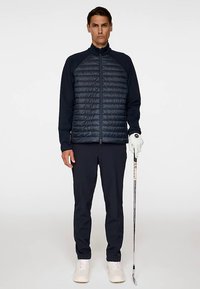 Laivastonsininen hybridi golf-takki tikatuilla edessä, pitkät hihat ja vetoketjusuljenta, yhdistetty tummiin housuihin ja valkoisiin kenkiin, kädessä golfmaila.