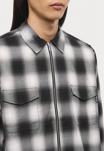 Black and white plaid shirt with a collar, featuring a front zipper, two chest pockets, and a textured fabric finish.