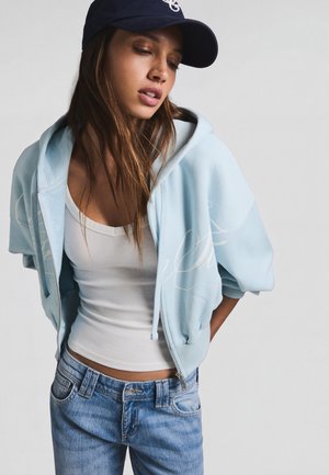 Jeune femme portant un sweat à capuche bleu clair zippé, un débardeur blanc, un jean bleu et une casquette foncée, regardant vers le bas avec les mains dans le dos.