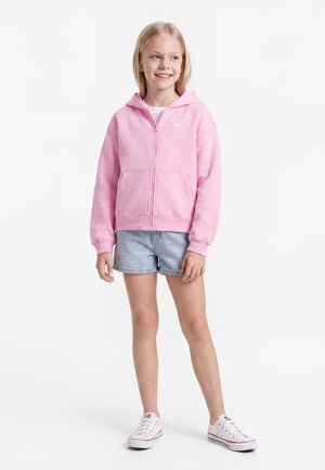 Enfant souriant debout portant un sweat à capuche rose zippé, un short en jean clair et des baskets blanches sur un fond blanc uni.