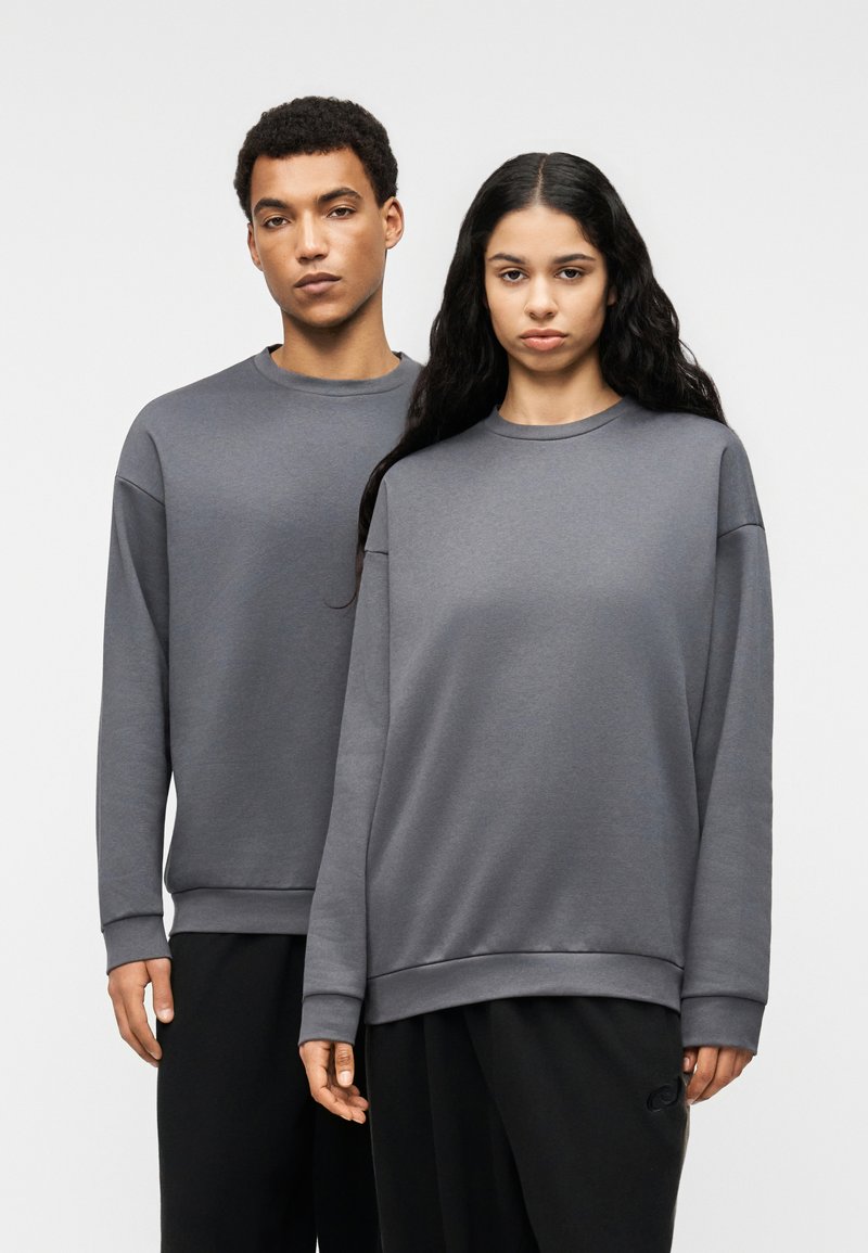 Δύο άτομα φορούν φαρδιές γκρι φούτερ με στρογγυλούς λαιμούς και ριμπ μανσέτες, συνδυασμένες με μαύρο παντελόνι. Το ύφασμα φαίνεται απαλό και λείο.