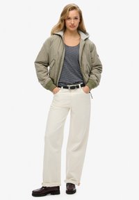 Chaqueta bomber caqui con forro de felpa gris, camiseta de rayas azul marino, pantalones anchos blancos y mocasines marrón oscuro con suela gruesa.