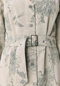 Abito beige con stampa floreale e di uccelli, caratterizzato da scollo a V, bottoni e cintura in vita. Realizzato in tessuto testurizzato con una finitura morbida.