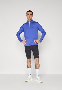 Blå langermet treningstopp med halv glidelås, sammen med svarte tettsittende shorts. Hvite joggesko med lysegrønne detaljer. Enkelt, funksjonelt design.