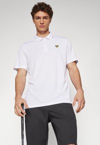 White polo shirt with a collar and short sleeves, featuring a small yellow logo on the left chest. Worn with dark shorts, holding a golf club.