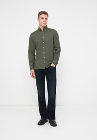 Camisa verde oliva de botones con mangas largas, combinada con vaqueros azul oscuro y zapatos negros. La camisa cuenta con un cuello y botones en el frente.