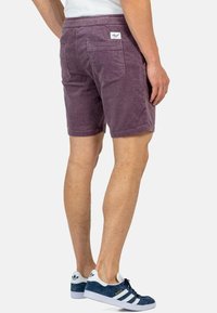 Pantalones cortos de pana morados con una textura suave, que cuentan con un bolsillo trasero de parche y una cinturilla elástica. Combinados con zapatillas azules.