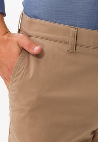 Pantaloni in misto cotone beige con una texture liscia, dotati di tasche laterali e un design a vita piatta. Cuciture visibili lungo i bordi.