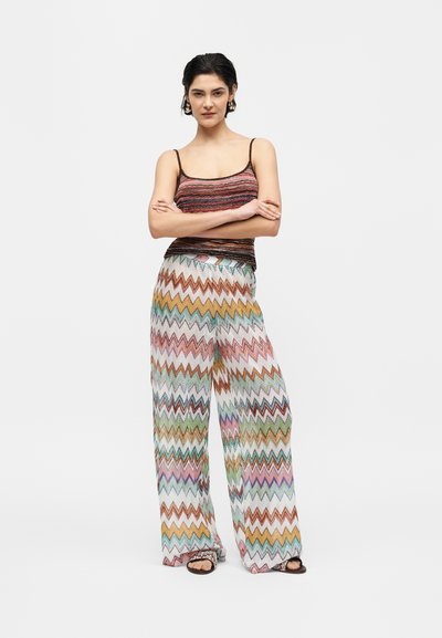 Γυναίκα που στέκεται με σταυρωμένα τα χέρια, φοράει αμάνικη ριγέ μπλούζα και πολύχρωμο παντελόνι με φαρδιά μπατζάκια και μαύρα σανδάλια.