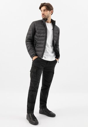 Mladý muž s rukama v kapsách, oblečený v černé prošívané bundě, bílém tričku, černých cargo kalhotách a černých teniskách, dívající se stranou.