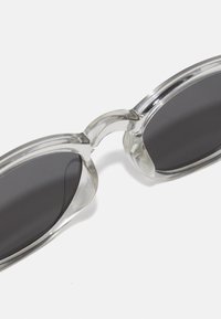 Klare Plastiksonnenbrille mit dunklen Gläsern, ovaler Form und breitem Rahmen. Glatte Textur mit einem leicht glänzenden Finish.