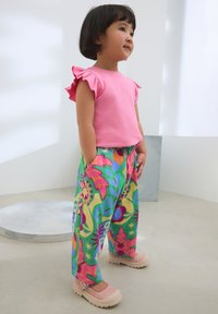 Pinkes Kurzarmoberteil mit Rüschendetails, kombiniert mit bunten, gemusterten Hosen mit Blumendesign. Hellrosa Slipper.