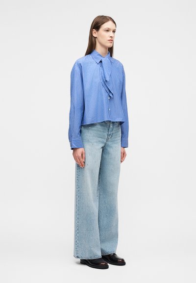 Femme debout portant un chemisier à manches longues rayé bleu avec un lien à l'avant et un jean bleu clair à jambes larges avec de petites décorations.