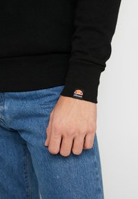Sudadera negra con puños acanalados, que presenta una pequeña etiqueta de logo multicolor. Combinada con unos vaqueros de mezclilla azules, mostrando un primer plano del área de la muñeca.
