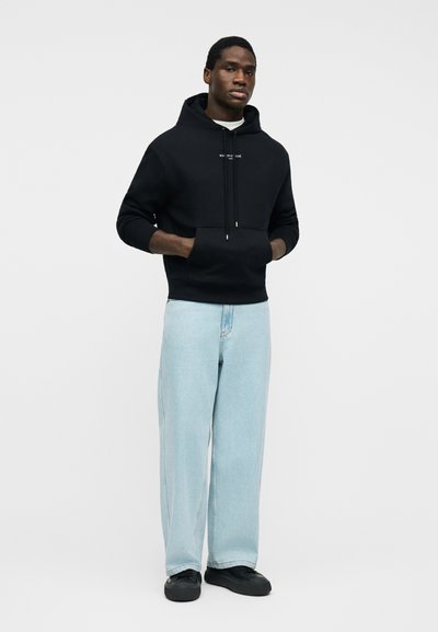 Άνδρας που φοράει μαύρο φούτερ με κουκούλα και μπροστινή τσέπη, καθώς και ανοιχτό μπλε τζιν με φαρδιά παντελόνια, στέκεται με τα χέρια στις τσέπες σε απλό φόντο.