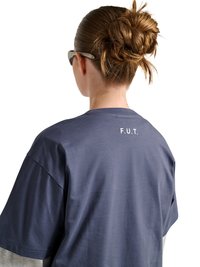 Kobieta z brązowymi włosami związanymi w niechlujny kok, nosząca okulary przeciwsłoneczne i granatowy luźny t-shirt z napisem "F.U.T." na górnej części pleców.