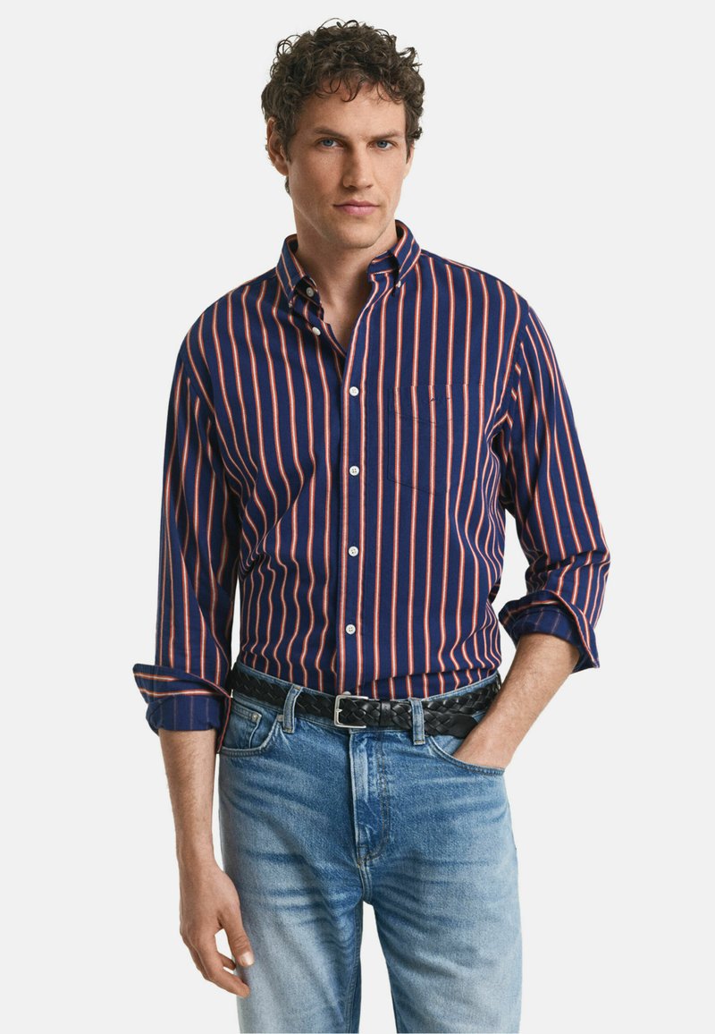 Chemise pour homme à manches longues, de couleur marine avec des rayures verticales orange ; en coton, avec un col, une poche poitrine unique, des manches retroussées, assortie à un jean en denim.