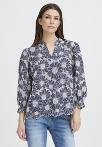 Blouse fleurie bleu marine avec un motif en dentelle blanche, un décolleté en V et des manches bouffantes. L'ourlet présente des bords festonnés. Portée avec un jean bleu clair.