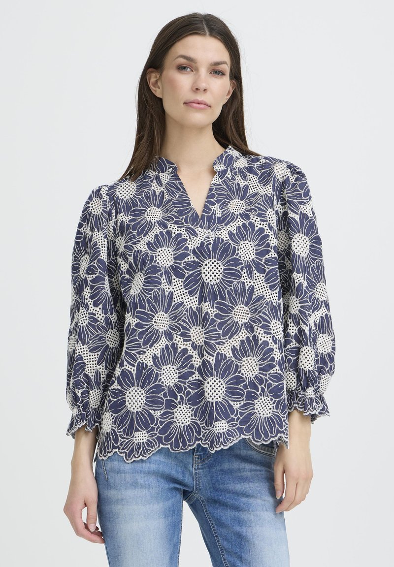 Blouse fleurie bleu marine avec un motif en dentelle blanche, un décolleté en V et des manches bouffantes. L'ourlet présente des bords festonnés. Portée avec un jean bleu clair.