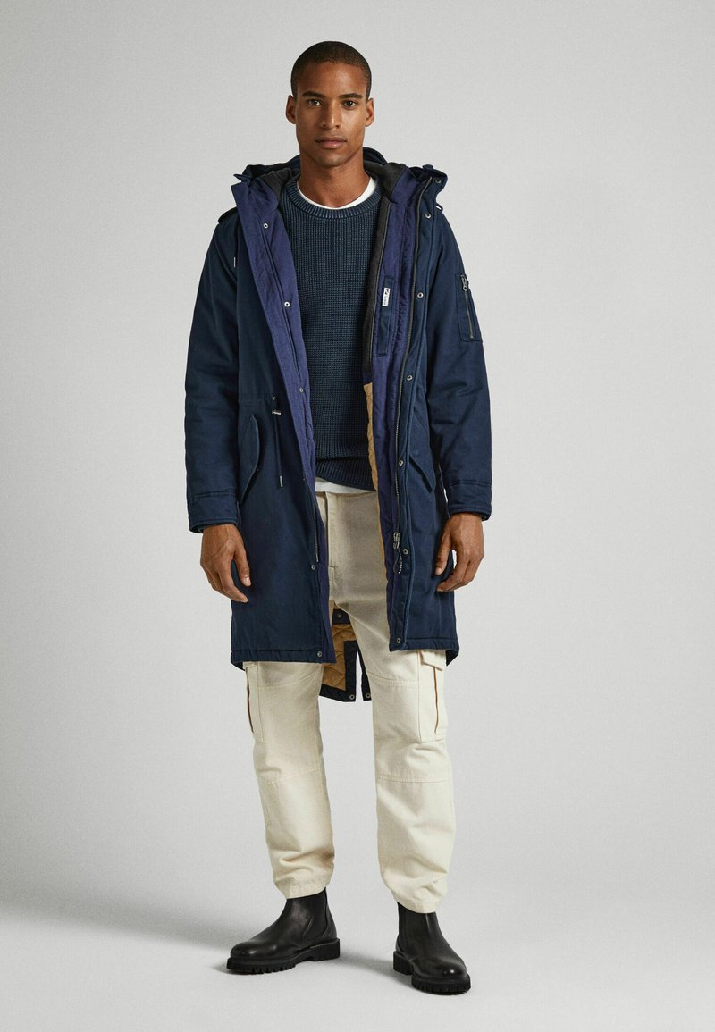 Giacca parka navy con cappuccio, caratterizzata da una chiusura con bottoni, due tasche laterali e un rivestimento imbottito. Abbinata a pantaloni cargo crema e stivali neri.