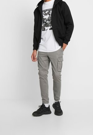 Homme portant un sweat à capuche noir zippé, un t-shirt blanc à imprimé camouflage, un pantalon cargo gris de style jogger, des chaussettes blanches et des baskets noires, debout une main dans la poche.