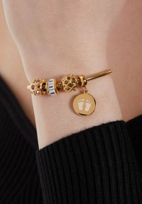 Gouden armband met ingewikkelde kraalontwerpen, roze stenen en een ronde charme met voetafdrukken. Geweven metallic afwerking overal.