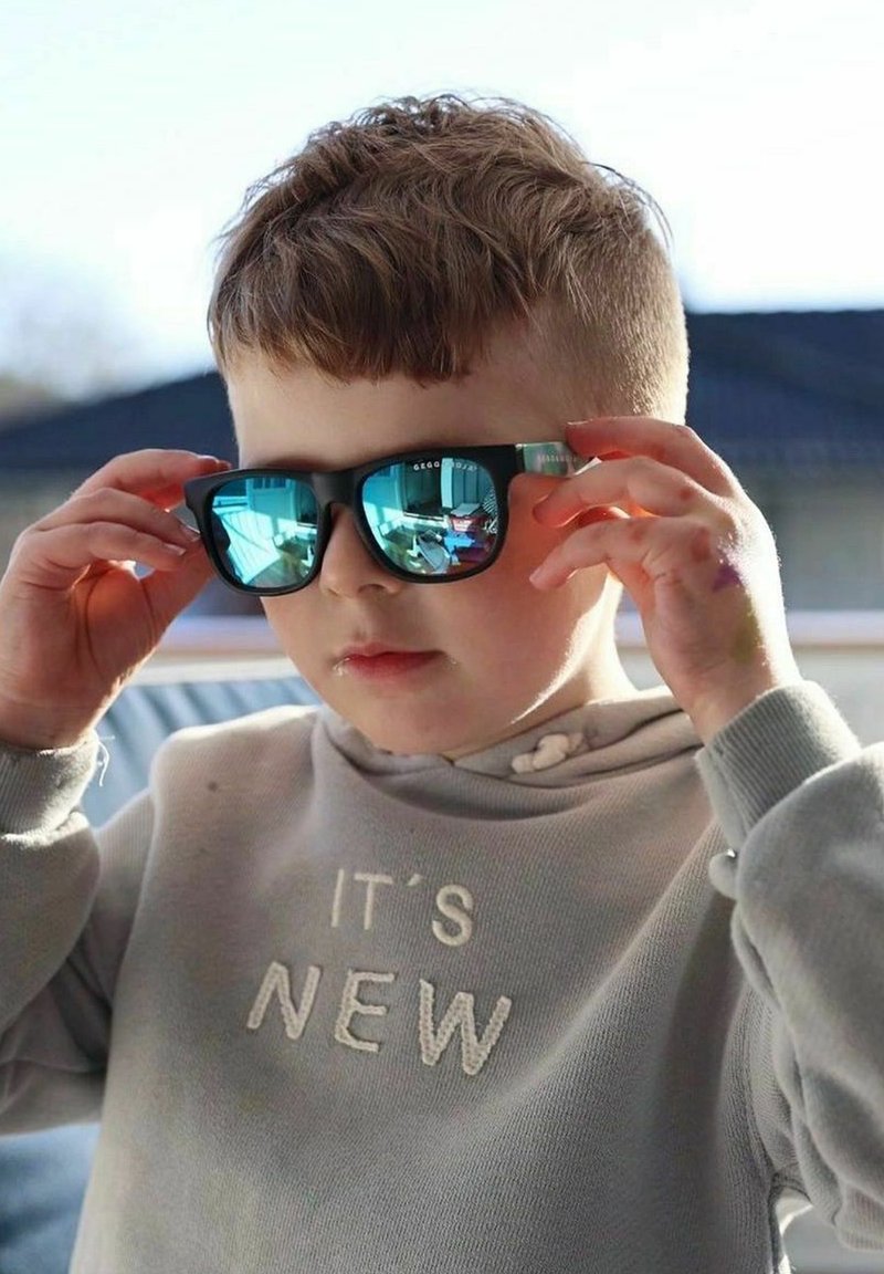 Svarta solglasögon med reflekterande blå linser, som bärs av ett barn. Barnet har på sig en ljusgrå huvtröja med vit text. Mjuk belysning framhäver detaljer.