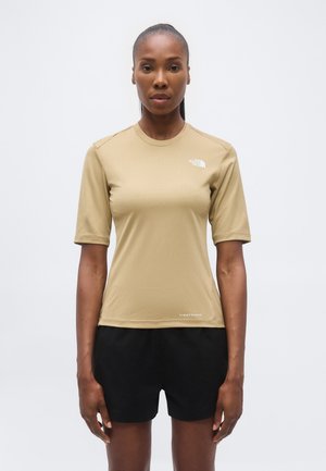 Kvinne som står vendt fremover, iført en beige kortermet topp og svarte shorts mot en ensfarget lys bakgrunn.