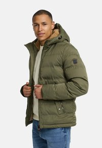 Zielona kurtka puchowa z podszewką ze sztucznego futra, zamkiem błyskawicznym i regulowanym kapturem. Posiada naszywkę na lewym ramieniu oraz dwie boczne kieszenie.