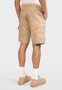 Mann trägt beige Cargo-Shorts mit mehreren Taschen auf der Rückseite und an den Seiten, weiße Socken und beige Turnschuhe, steht vor einem weißen Hintergrund.