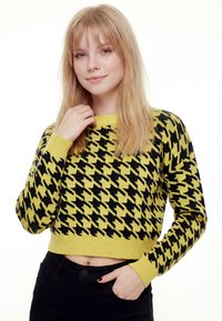 Čierno-žltý krátky sveter so výrazným vzorom psieho zubov, pletenou textúrou a rebrovanými manžetami a lemom. Kombinovaný s čiernymi nohavicami.