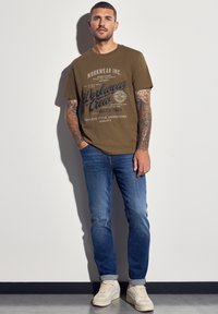 Olivově zelené bavlněné tričko s modrým grafickým textem, spojené s světle modrými džíny. Model má na sobě béžovou obuv. Pozadí je jednobarevné.