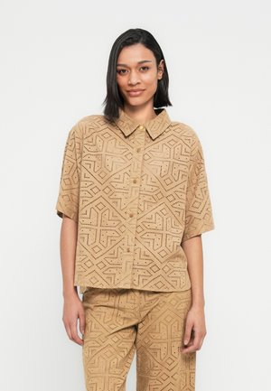Chemise à manches courtes couleur sable avec un motif géométrique découpé, col à boutons et fermeture à boutons sur le devant. Tissu texturé avec une coupe décontractée.